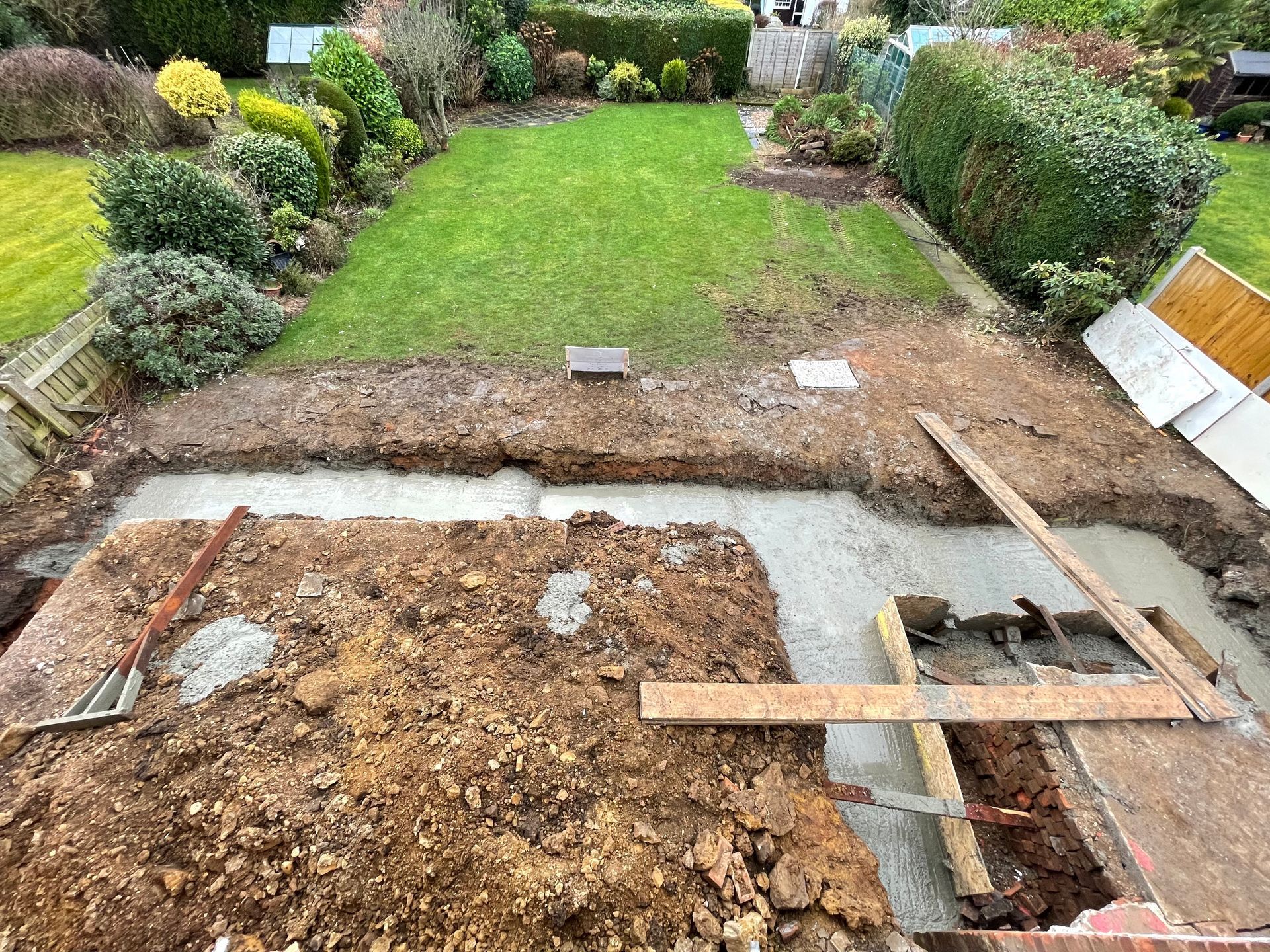 Backyard with construction: excavated soil, concrete foundation, grass lawn, and garden beds.