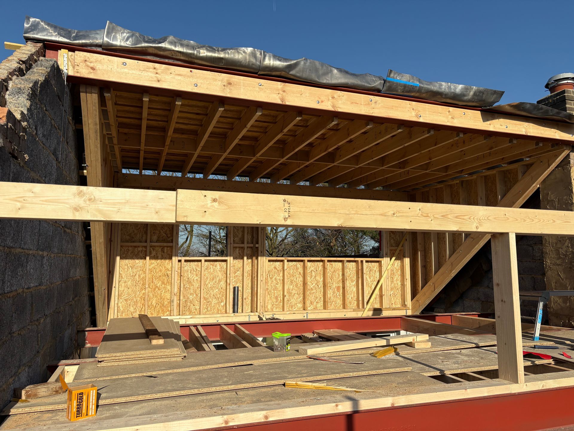 Construction site with wooden frame structure, possibly a roof extension. Brown wood, clear sky.