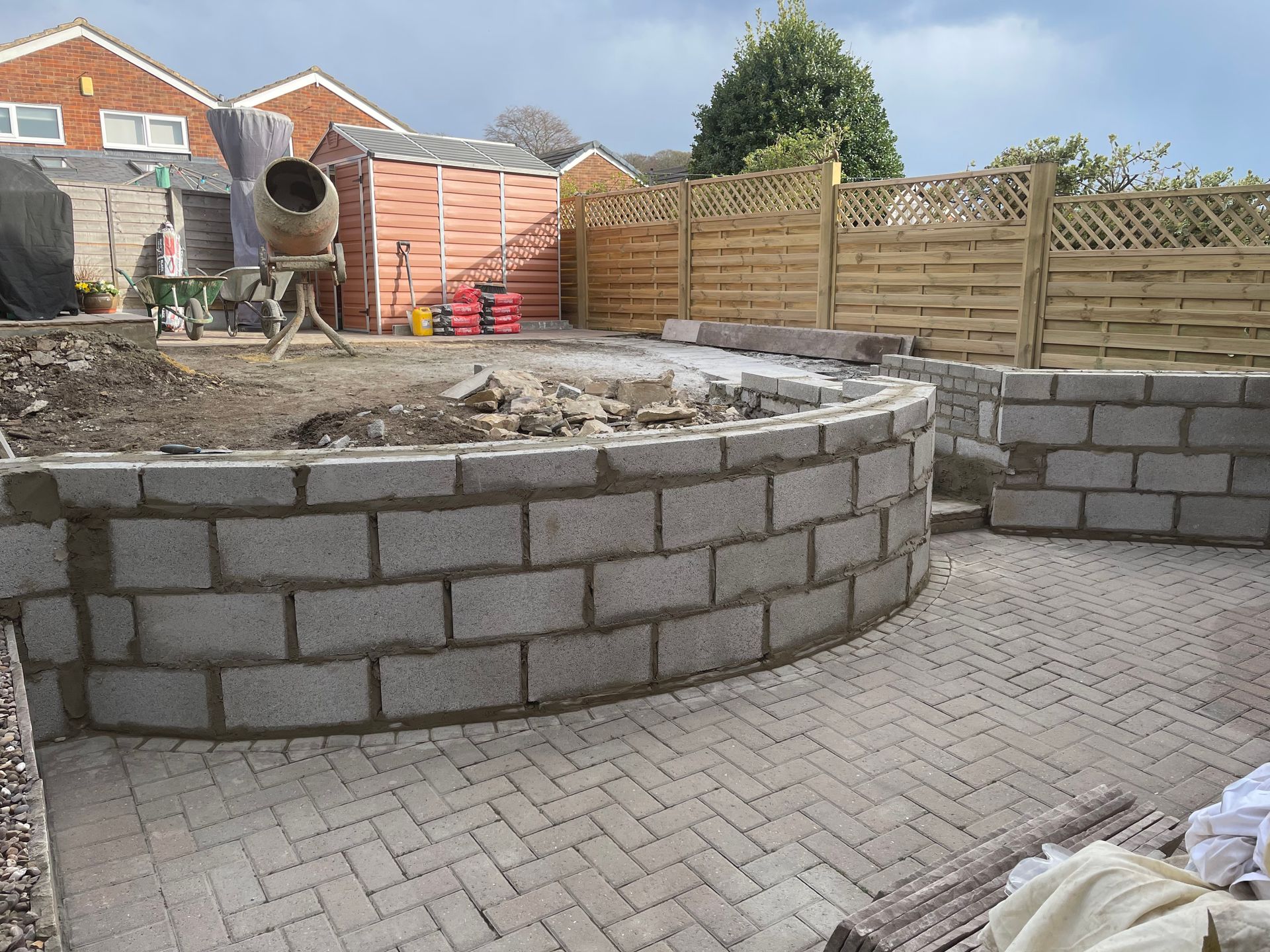 A curved brick retaining wall under construction in a backyard, with a paved patio and a shed in the background.
