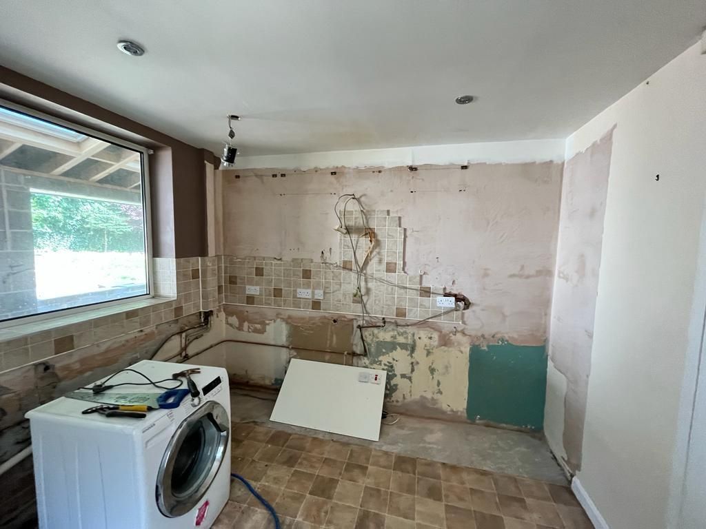 Kitchen under renovation: bare walls, washer, electrical wiring visible, old tile, and open window.