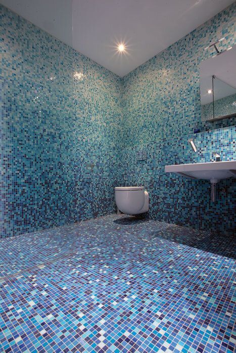 Blue mosaic-tiled bathroom. Wall-mounted toilet and sink. Light fixtures reflect off the glossy surfaces.