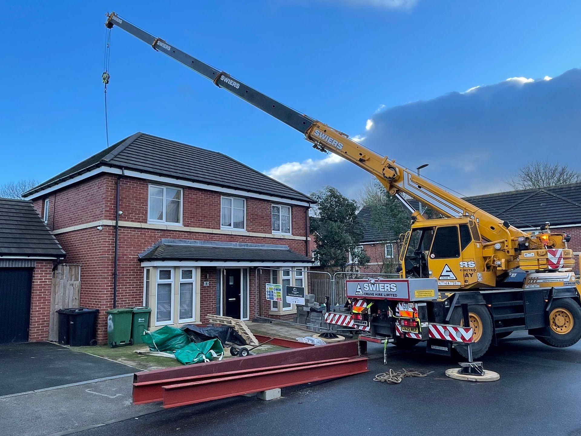Yellow crane lifting a metal beam in front of a two-story brick house. Construction site on the driveway.