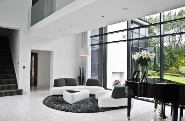 Modern living room with grand piano, white sofas on a black rug, and large windows overlooking a garden.