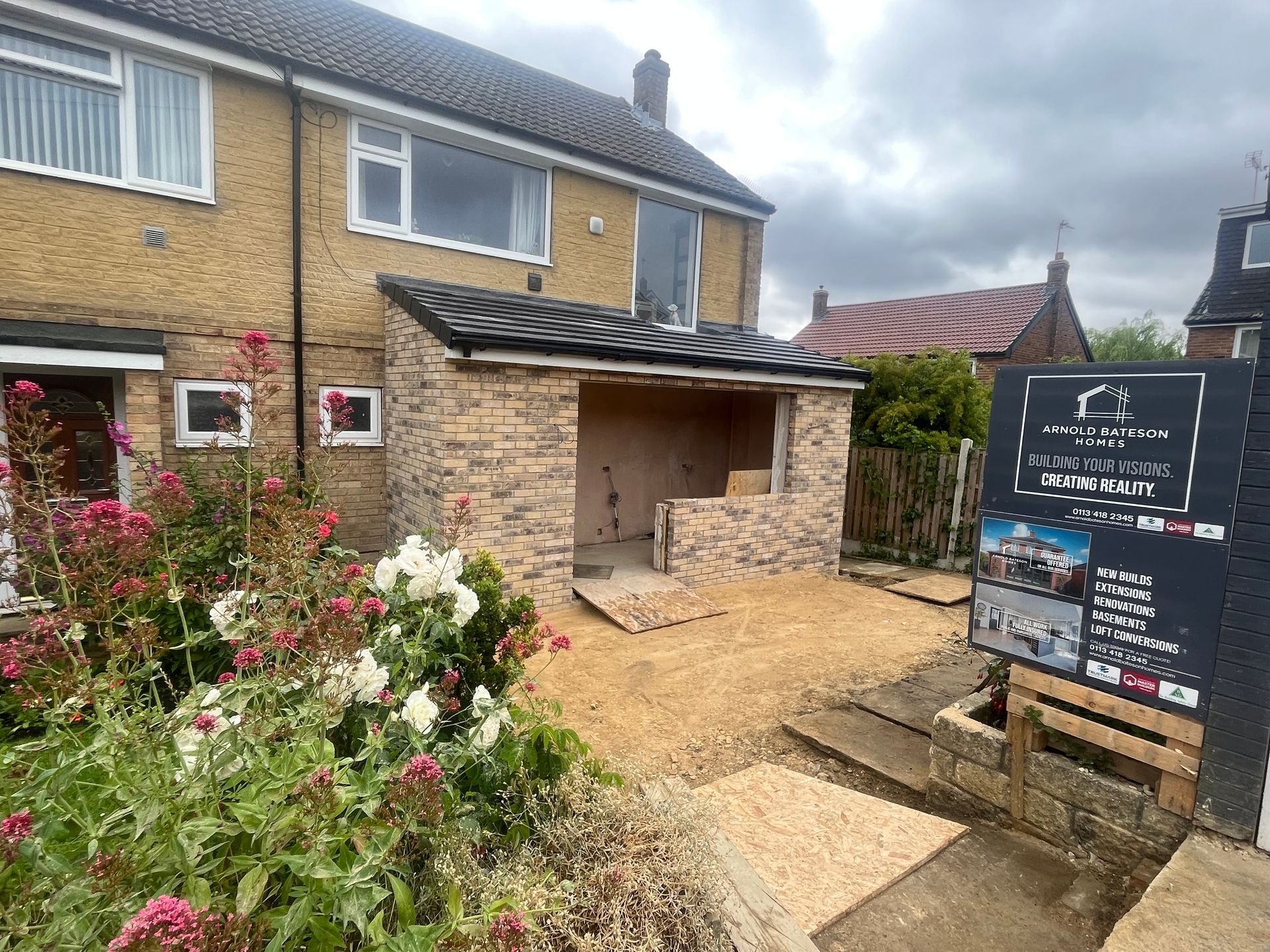 Construction of a tan stone extension on a brick house, gravel path, sign on the right.