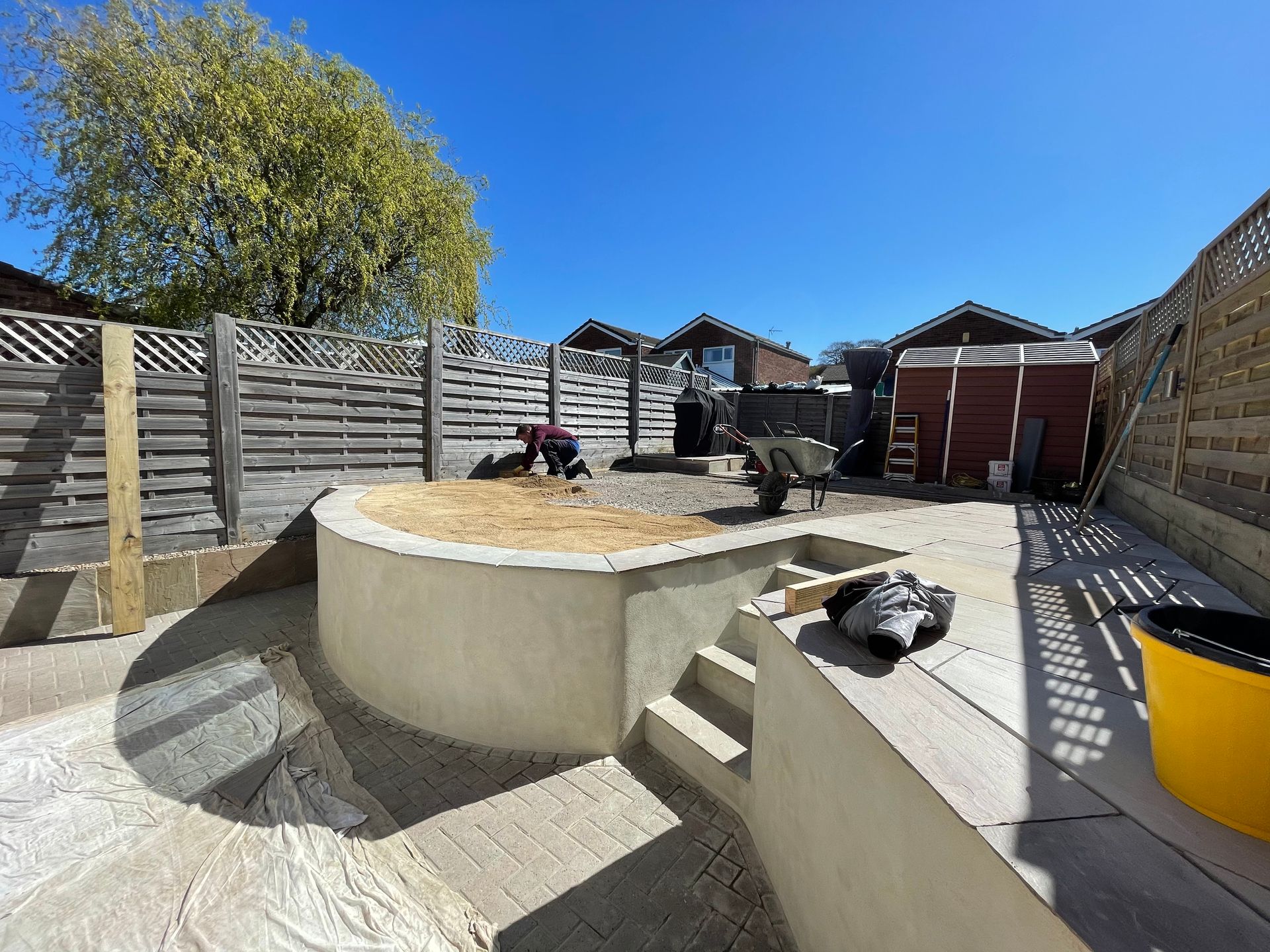 A sunny backyard under construction, with a stone patio, retaining wall, and person working near a shed.