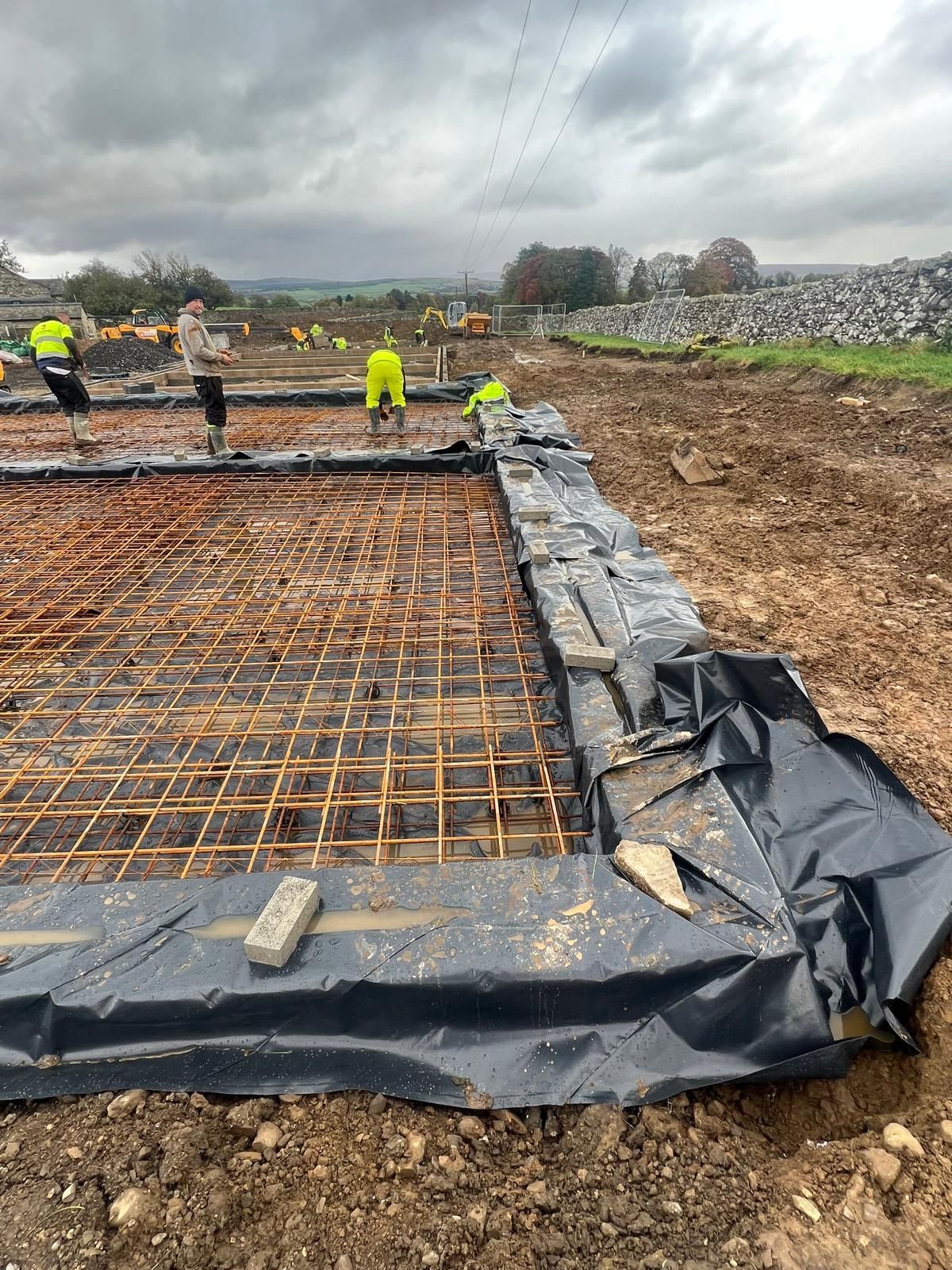 Construction site: Workers laying rebar grid, bordered by black plastic, for concrete foundation.