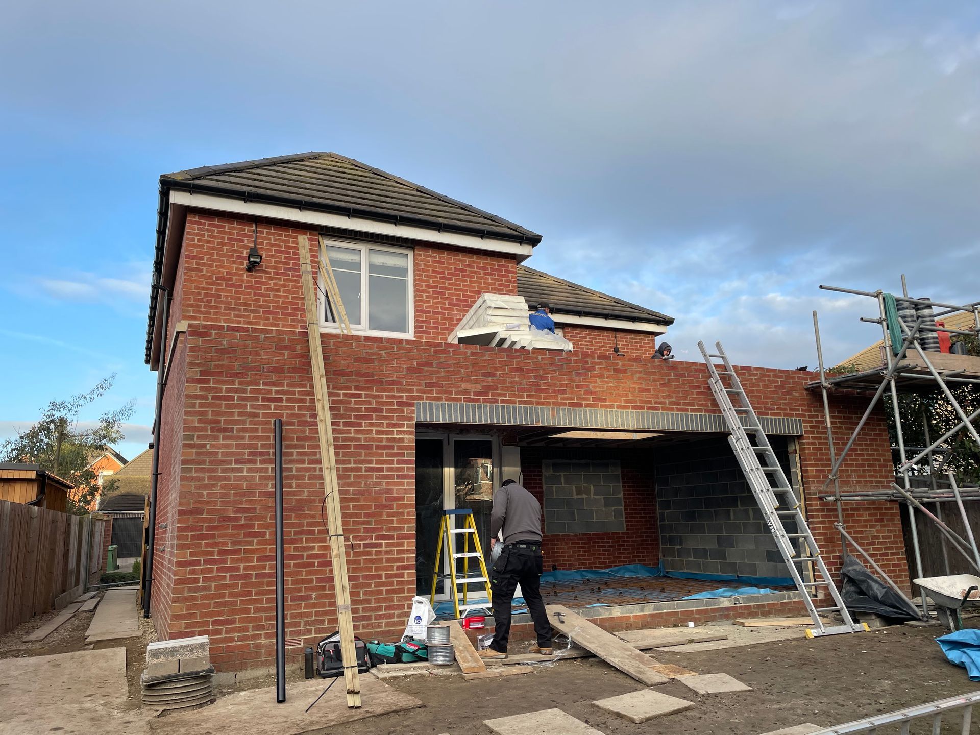 Brick home under construction; worker stands at doorway, ladder nearby, scaffolding to the right.