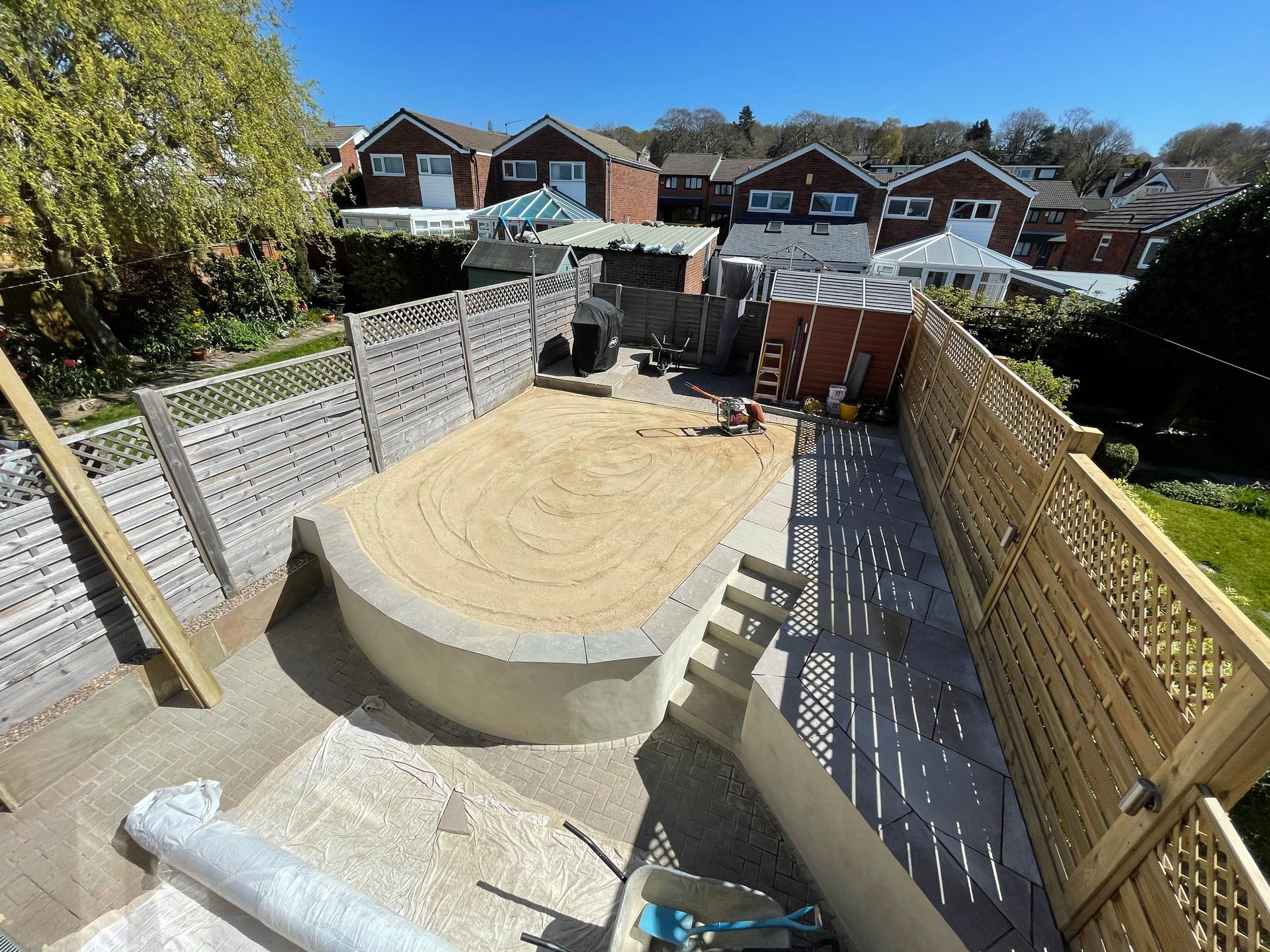Backyard under construction: Sand-filled space, curved wall, fence. Workers near shed and surrounding houses on a sunny day.