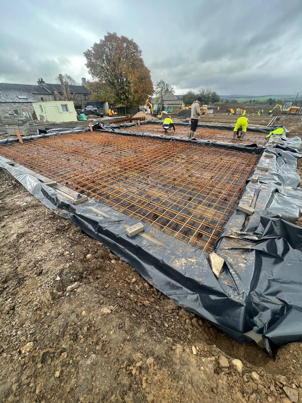 Construction site: workers around a rebar grid laid over a black membrane, ready for concrete, surrounded by mud.