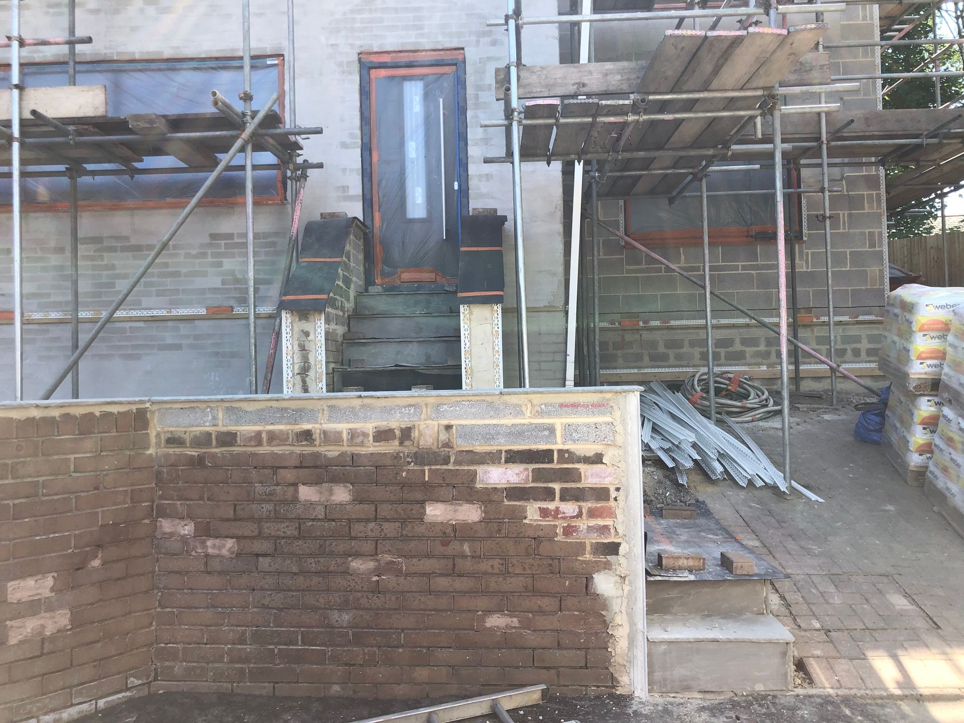 Exterior of a house under construction with brickwork, scaffolding, and a front door visible.