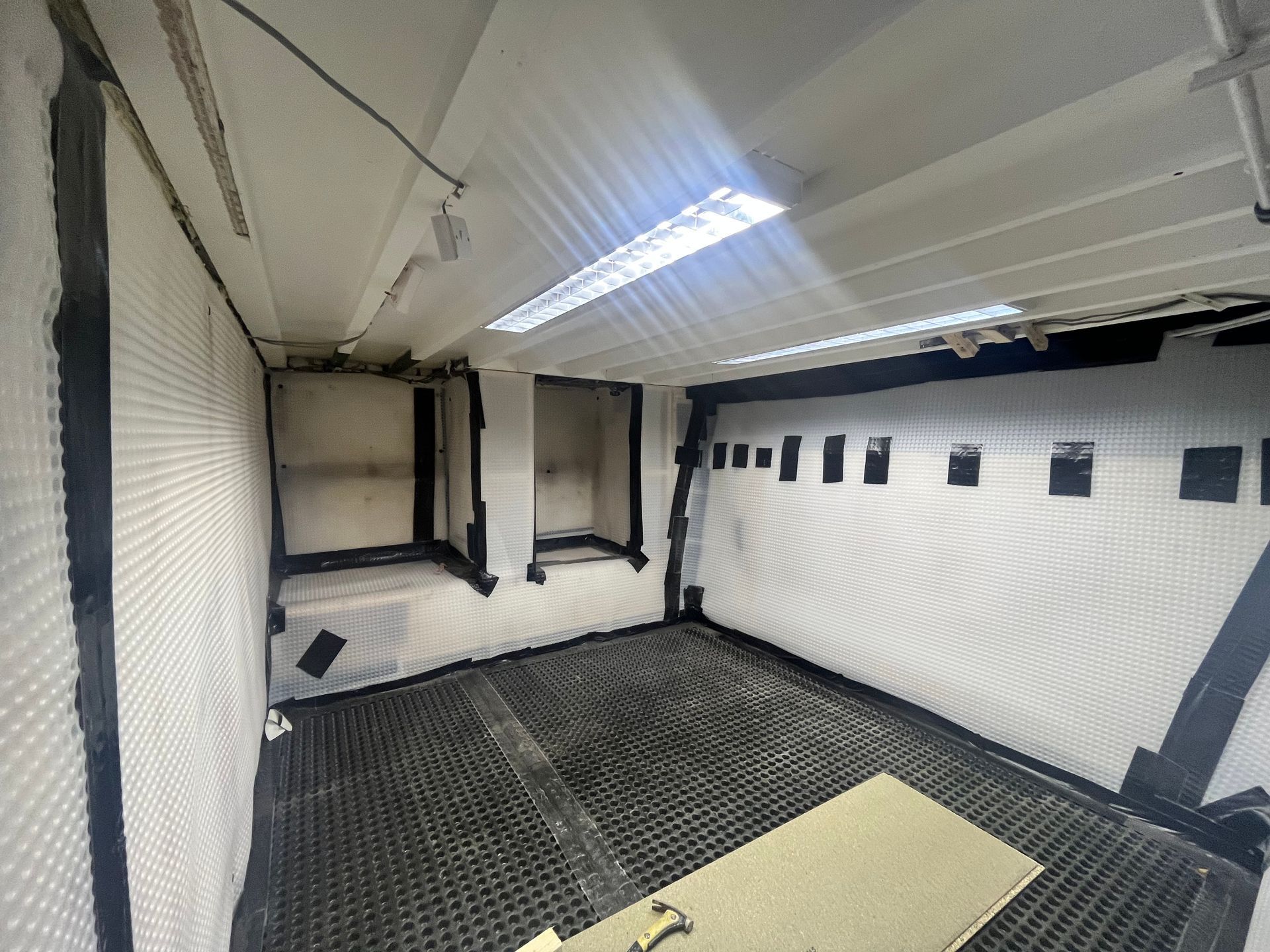 Interior of a white room with black grates and tape. Fluorescent lights overhead, with multiple small windows