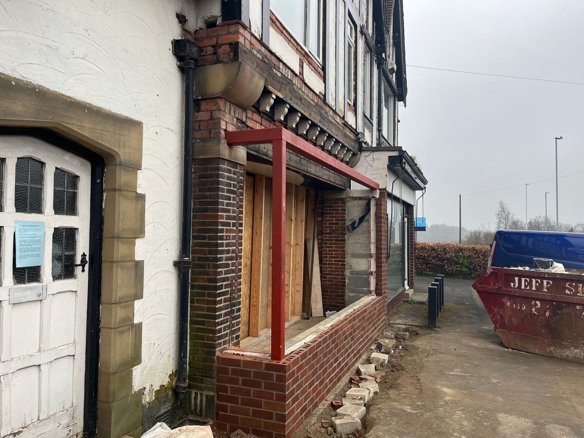 Building exterior under renovation; brickwork, red metal frame, white door, and a skip.