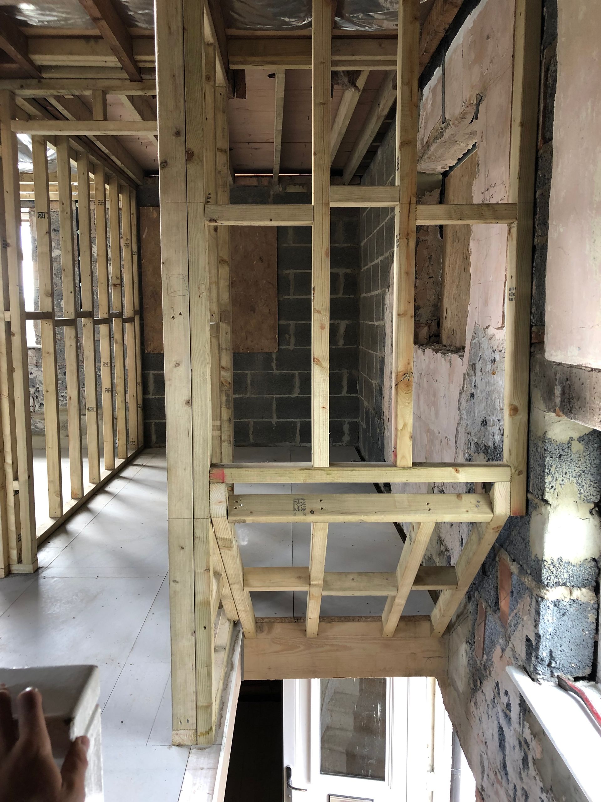 Interior view of a building under construction, with wooden framing. A hallway and stairwell are visible.