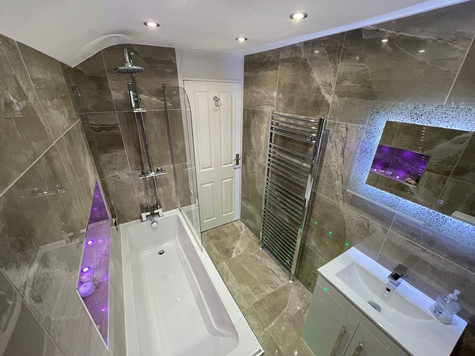 Modern bathroom with gray tile, tub, shower, heated towel rack, sink, and illuminated mirror.