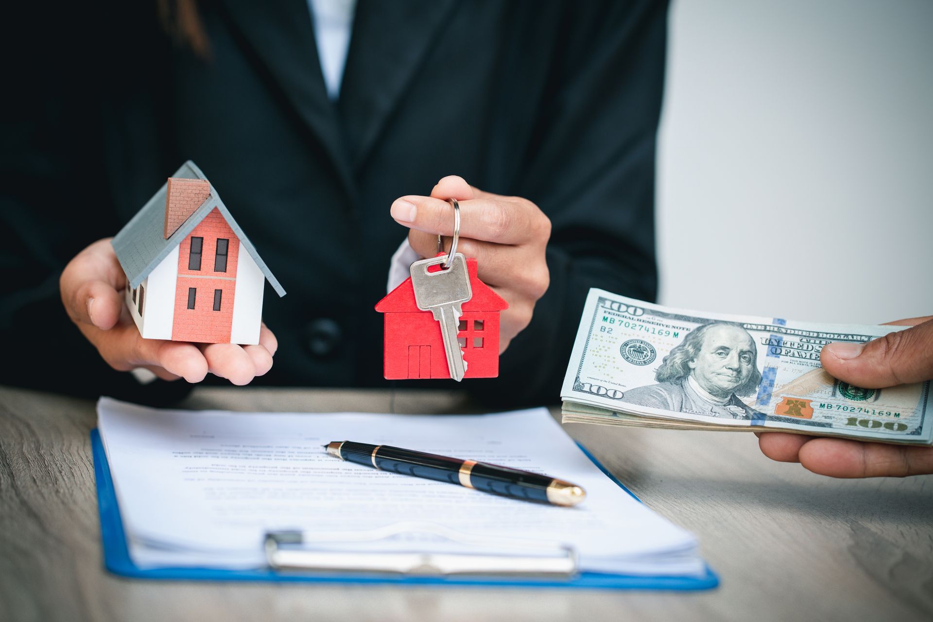 Person holding house keys and model house, receiving cash over a document and pen.