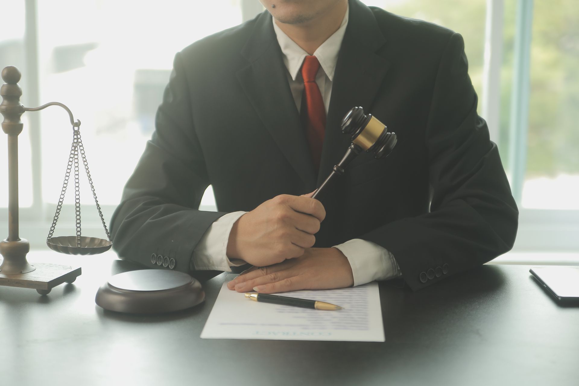Lawyer in black suit holding a gavel over documents, scales of justice and pen on desk.