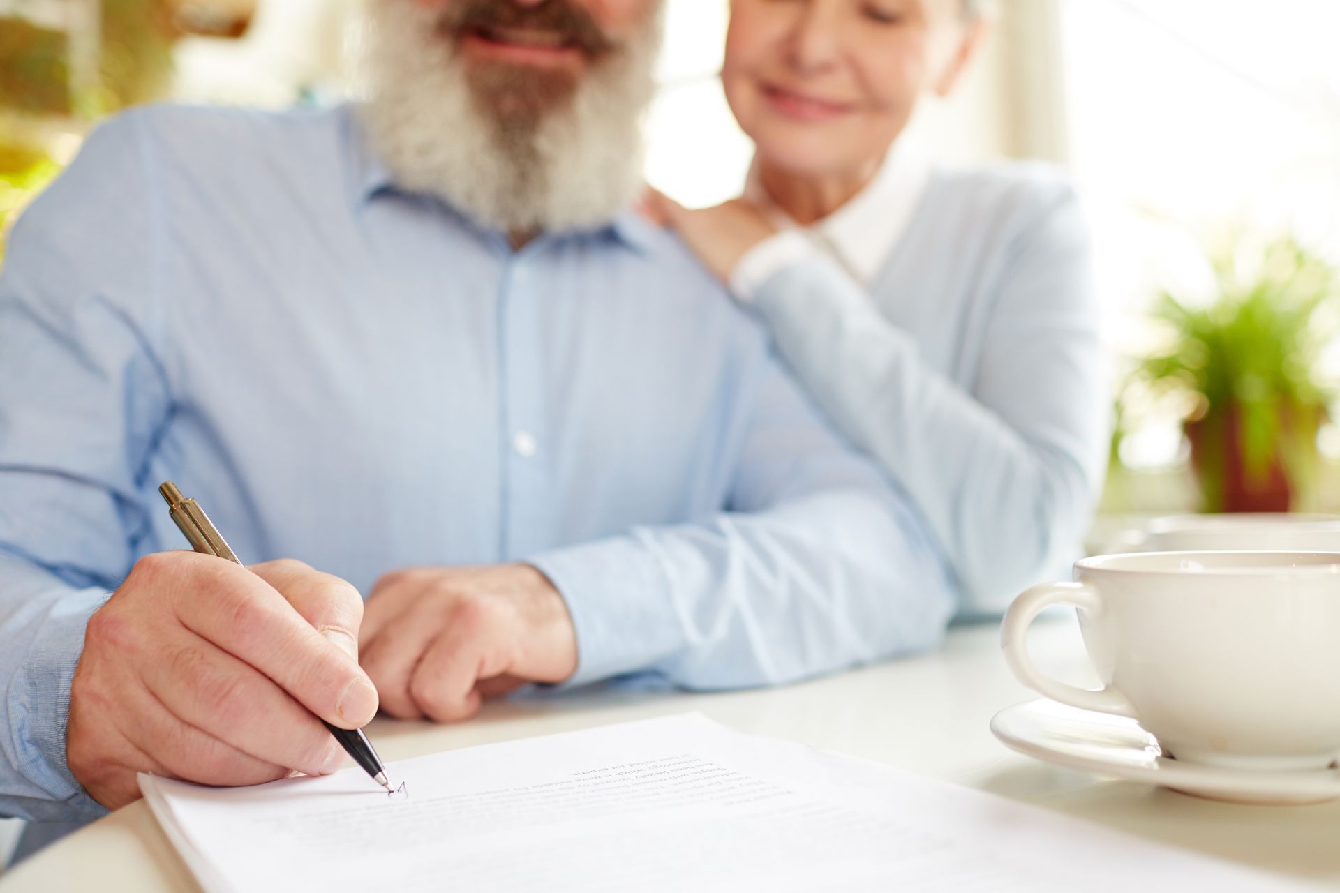 Man writing at a table, woman smiles behind him. Coffee cup on table, bright setting.