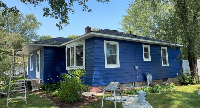 Blue house with white trim, set in a yard with grass and trees under a clear blue sky.