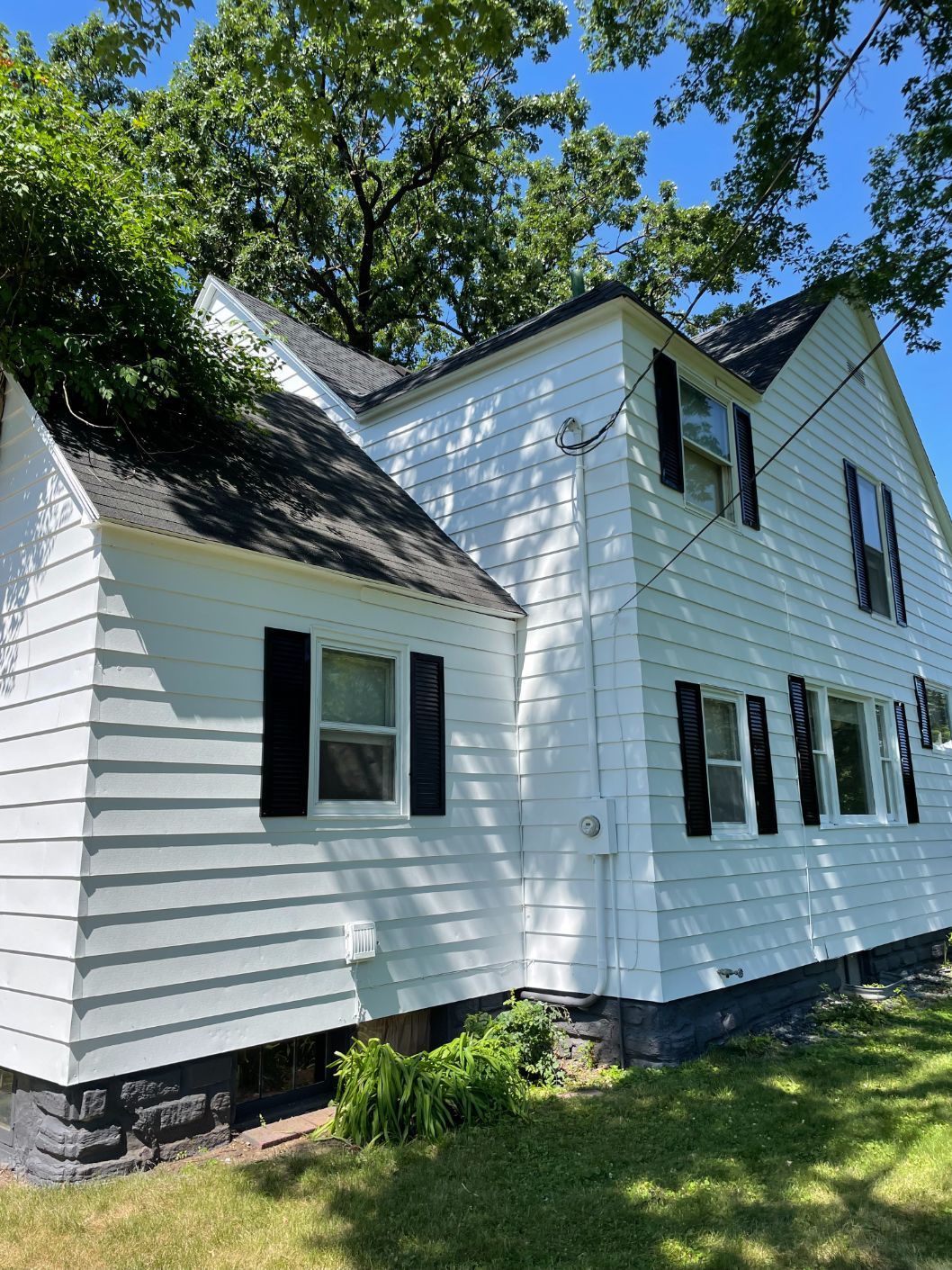 White two-story house with black shutters, roof partially covered by tree branches, blue sky.