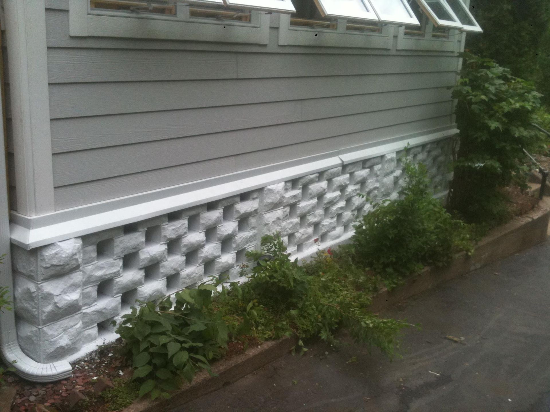 Grey house with patterned concrete block foundation, bushes, and a downspout.
