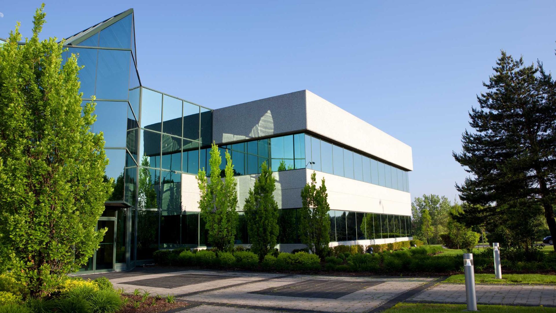 Modern glass and white building with surrounding greenery under a blue sky.