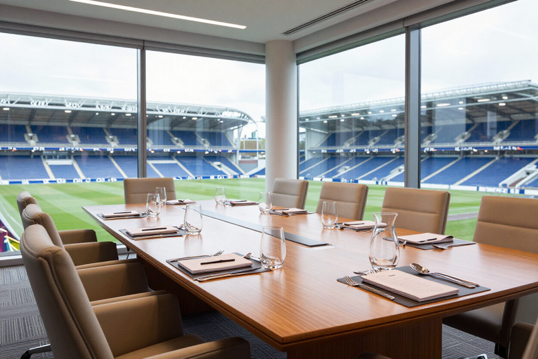Modern conference room with a wooden table and beige chairs overlooking a blue stadium through large windows