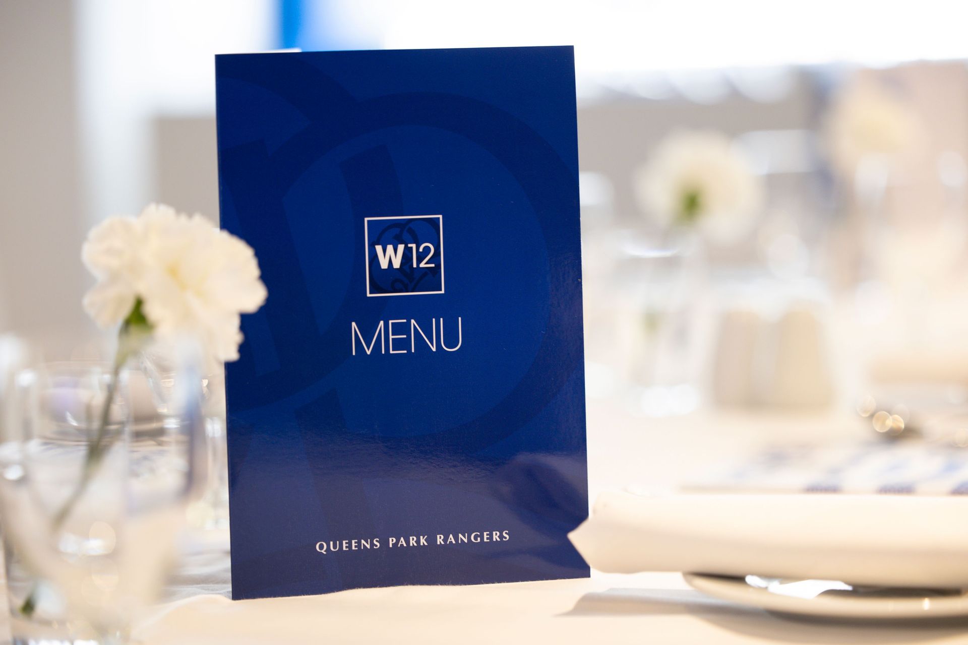 Blue menu card with “W12 MENU” on a set dining table, with flowers and tableware in the background