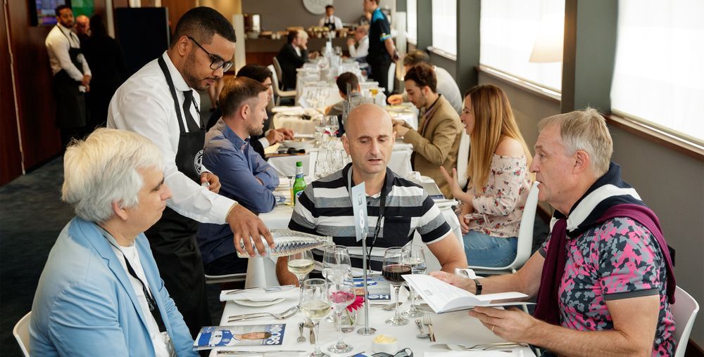 People seated at a dining table in a bright restaurant, chatting over drinks and menus.