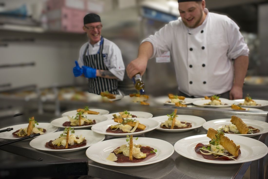 Chefs plating gourmet dishes in a busy kitchen, with white plates arranged on a counter.