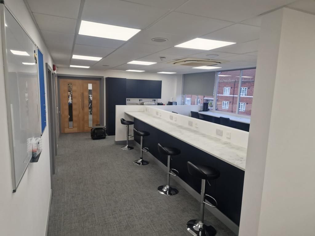 Empty office break room with long counter, black stools, gray carpet, and windows along the right wall.