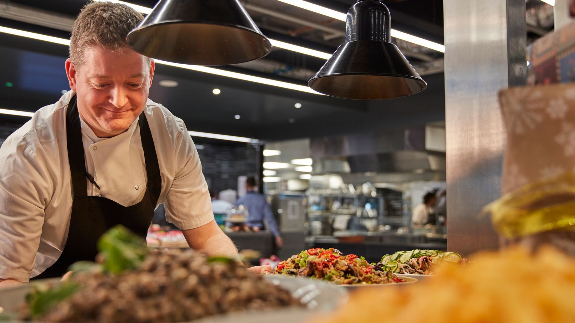 Chef plating food in a busy restaurant kitchen under hanging heat lamps.