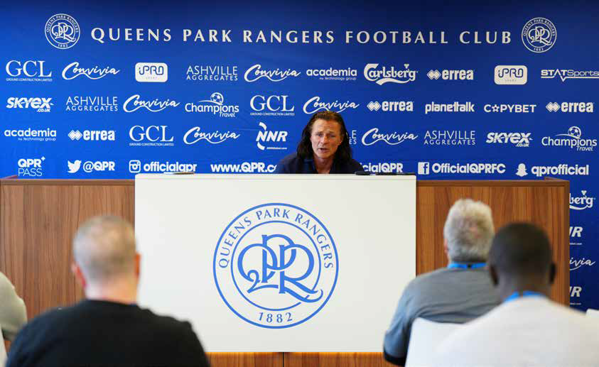 Queens Park Rangers press conference with speaker at podium before blue sponsor backdrop