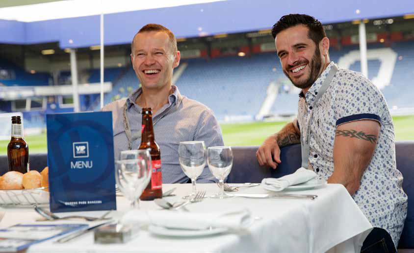 Two men smiling at a table with drinks in a stadium restaurant, a menu displayed in front