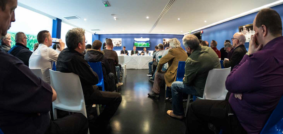 People seated in a conference room watching a presentation on a large screen at the front