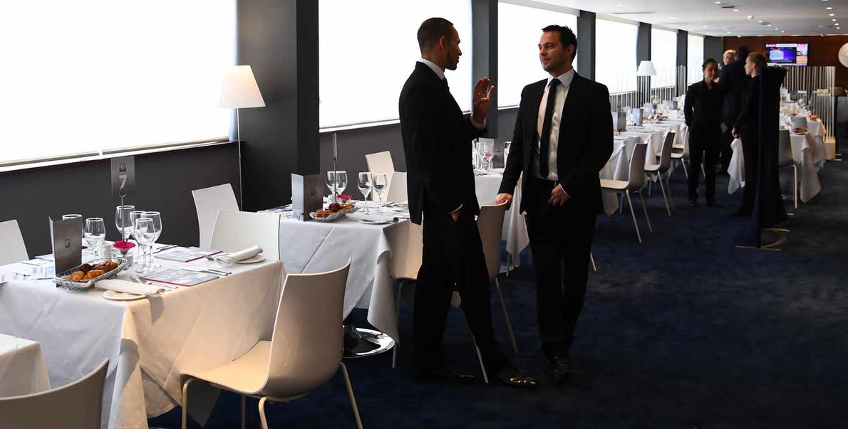 Conference hallway with two men talking beside white-clothed tables and chairs; people gather in the background.