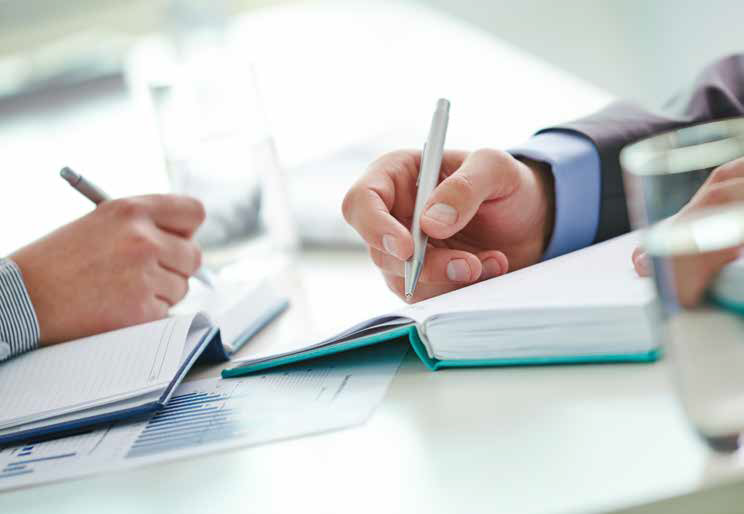 Two people writing in open notebooks at a bright table with pens and coffee cups