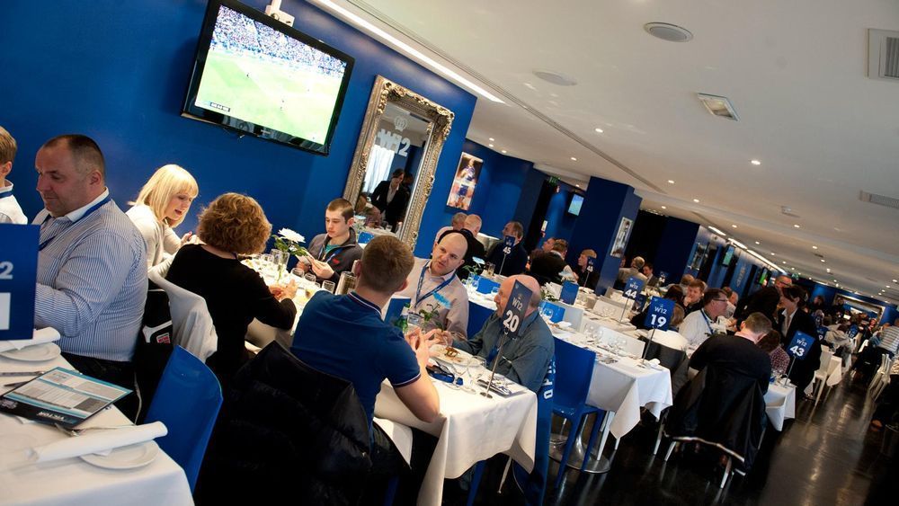 People dining at a blue-and-white banquet hall with large TV screens showing a sports game.