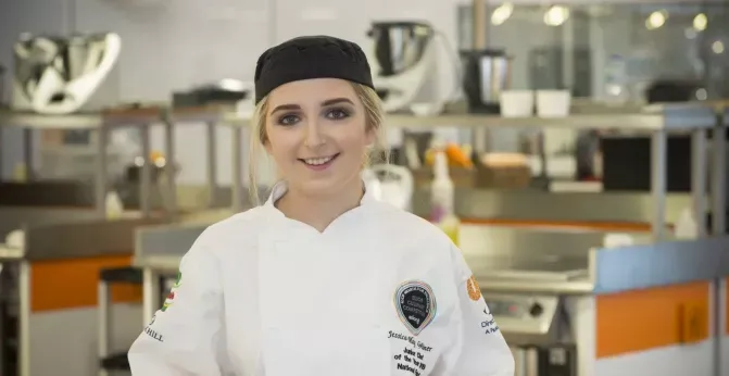 Chef in white uniform and black cap standing in a professional kitchen, smiling at the camera
