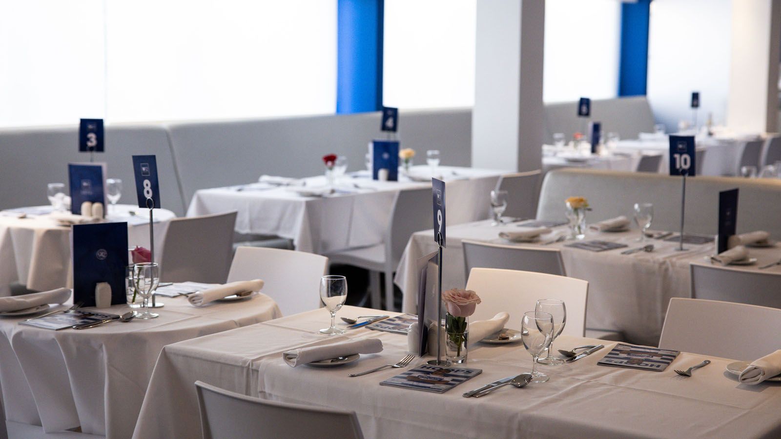Elegant dining tables set with white linens, glassware, and blue table numbers in a bright restaurant interior