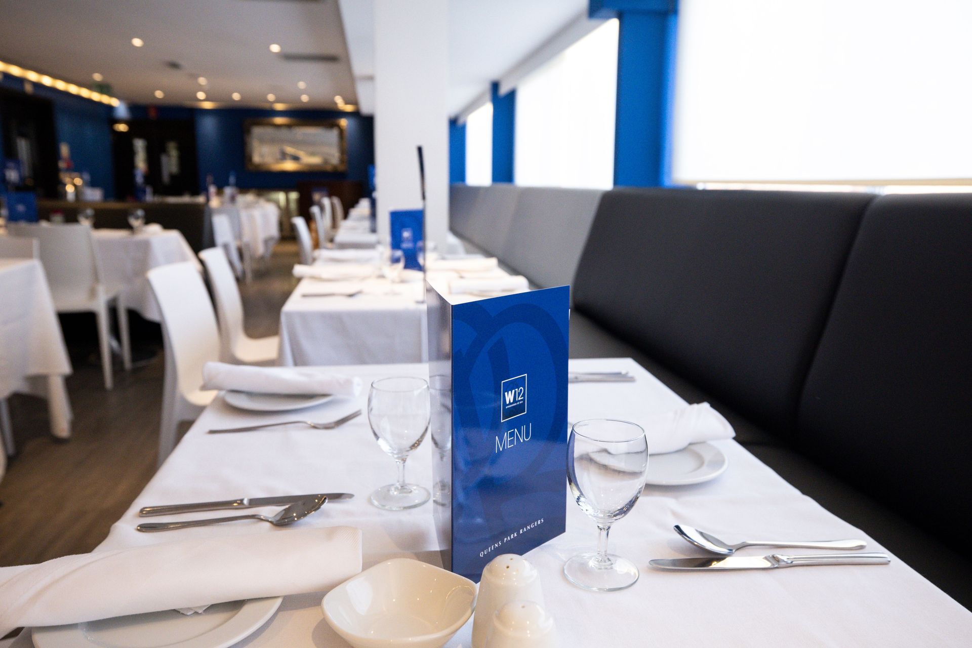 Set restaurant table with white linens, black bench seating, and blue menu card in a modern dining room