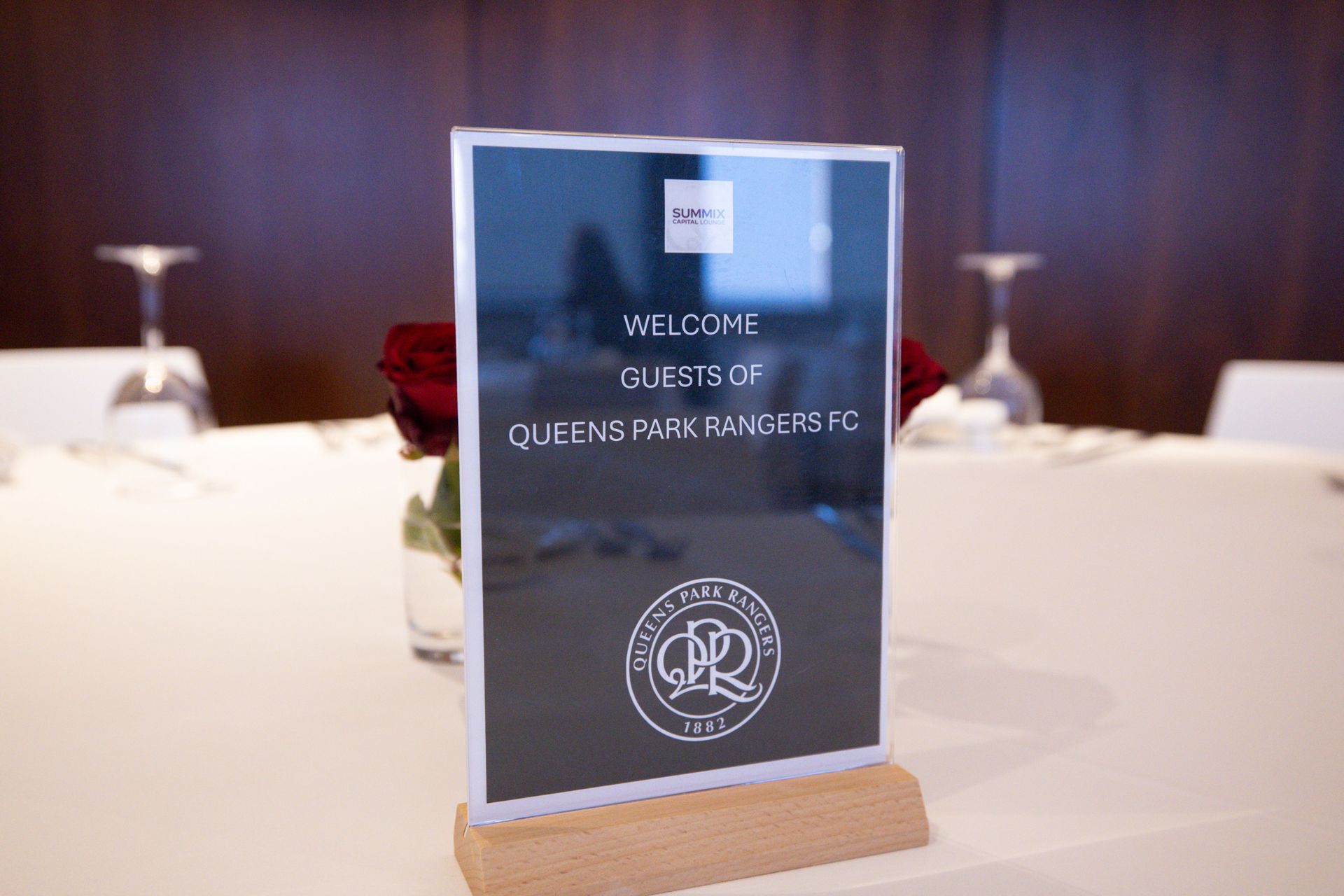 A framed welcome sign on a wooden stand at a formal event table with white tablecloths and wine glasses.