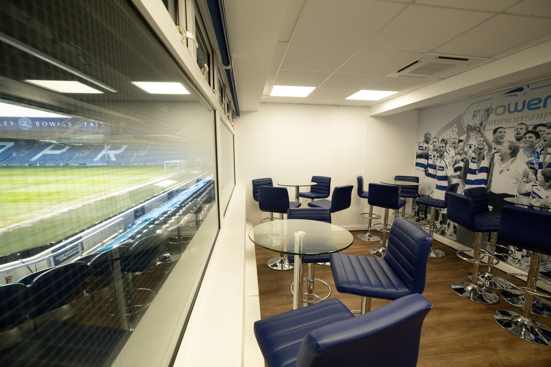 Stadium lounge with blue chairs, glass table, and a view of the soccer field through a large window