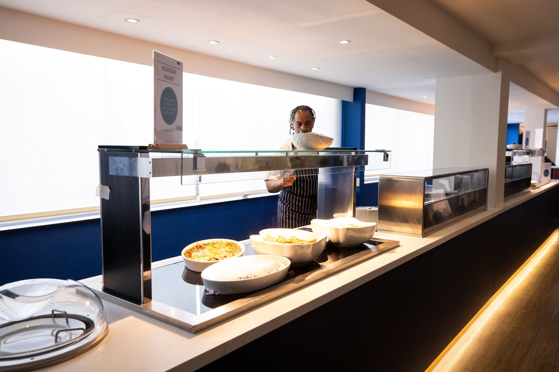 Restaurant buffet counter with covered dishes and a staff member behind glass sneeze guards