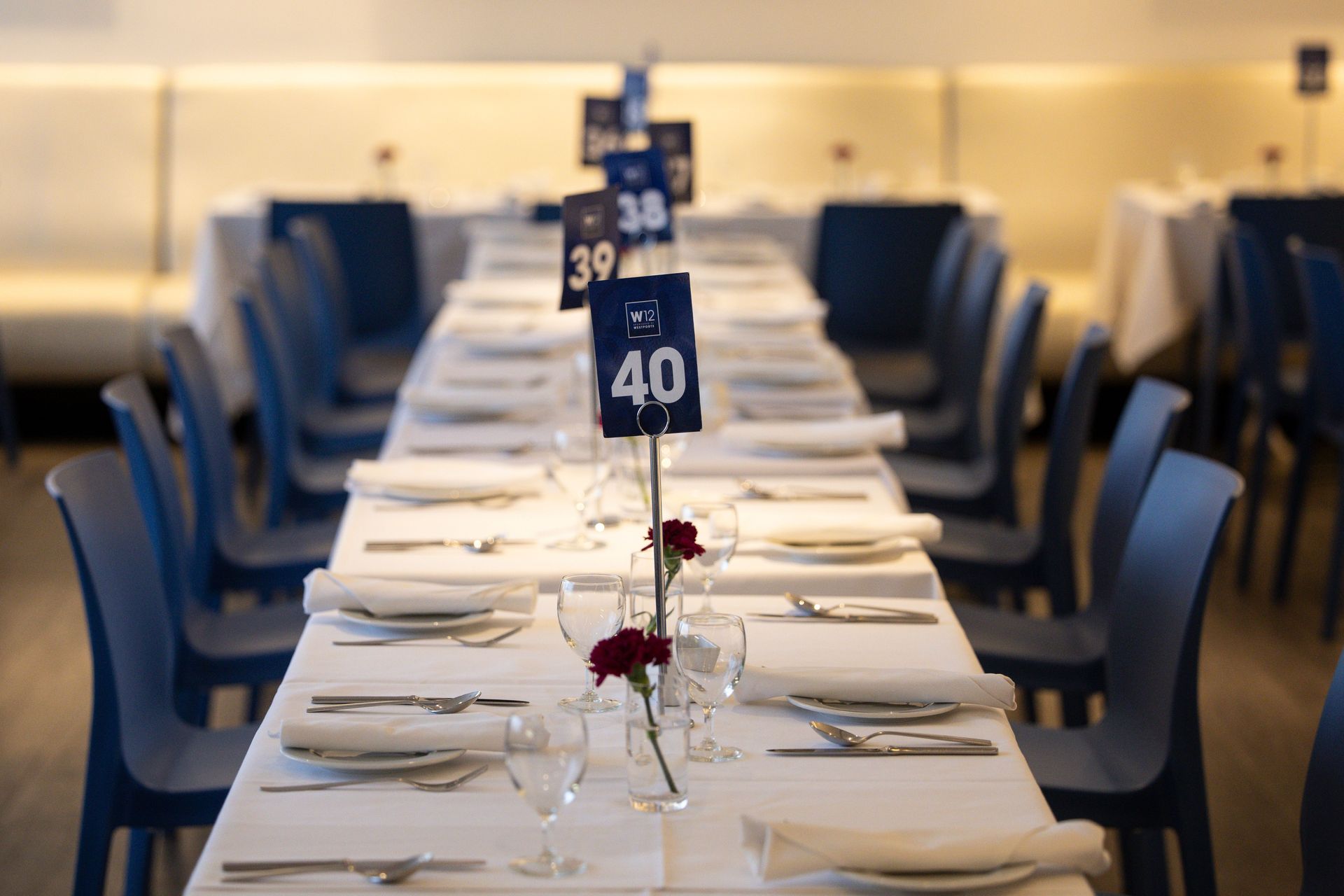 Elegant banquet table set with white linens, folded napkins, wine glasses, and numbered centerpieces in a blue-lit room