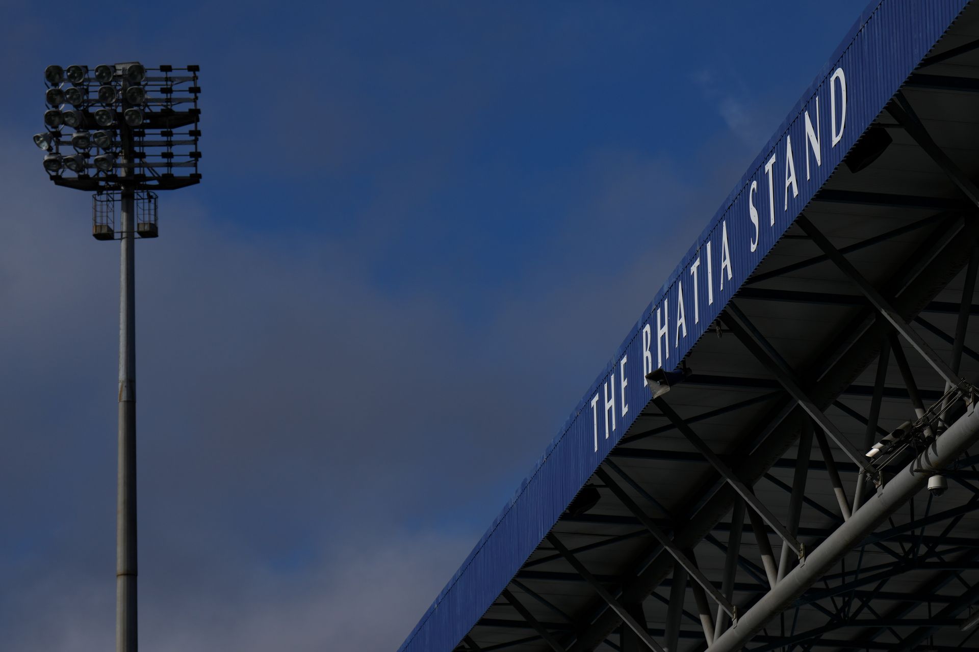 Stadium corner with blue roof and floodlight tower against a cloudy evening sky