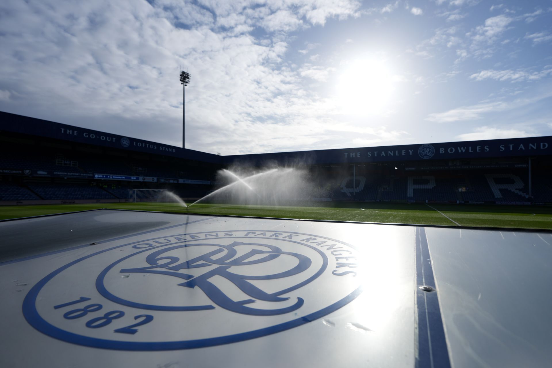 Rangers FC logo on a shiny surface in a stadium, with sprinklers spraying under bright sunlight