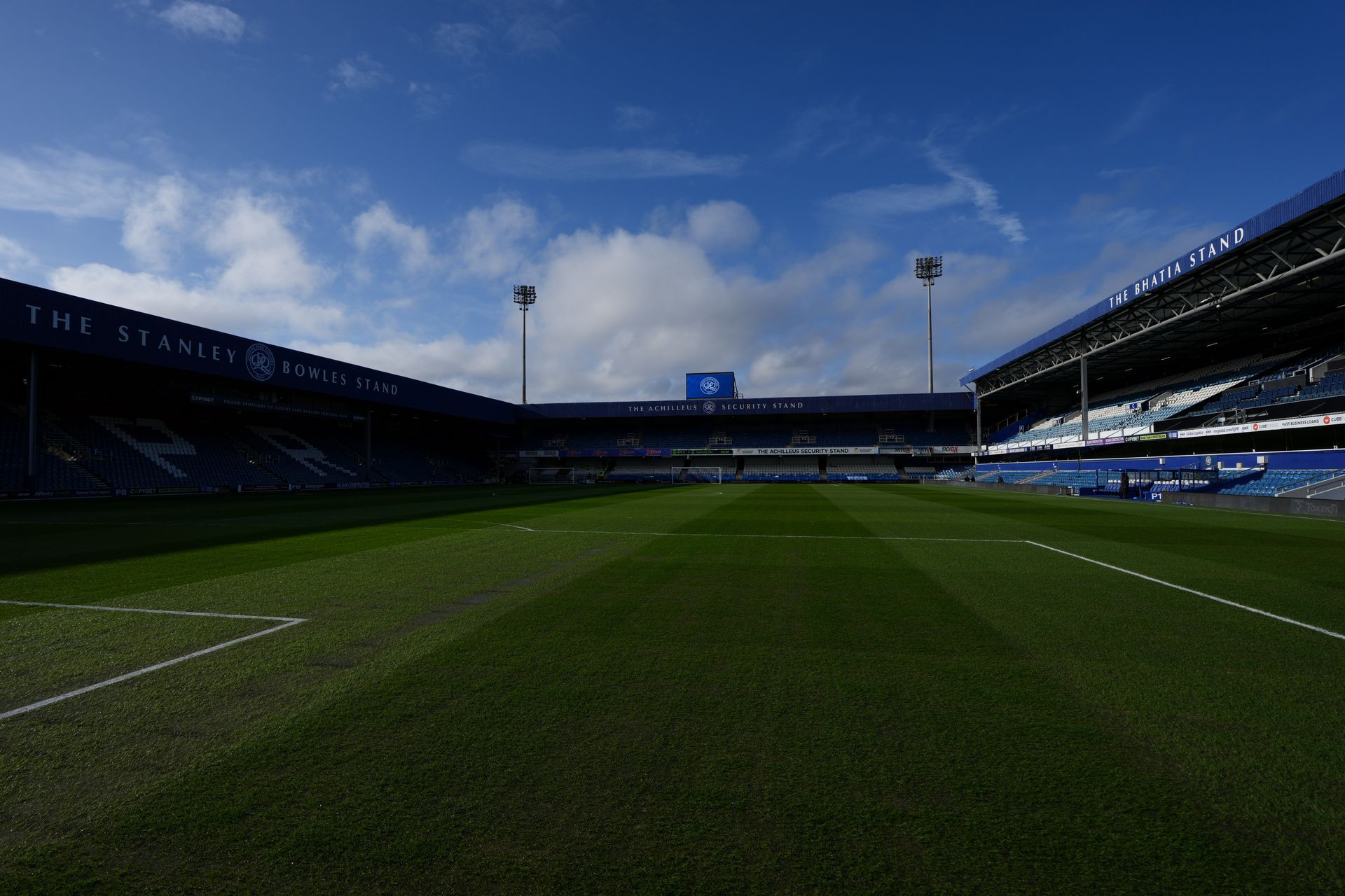 Empty soccer stadium under a blue sky with green pitch and stands in shadow