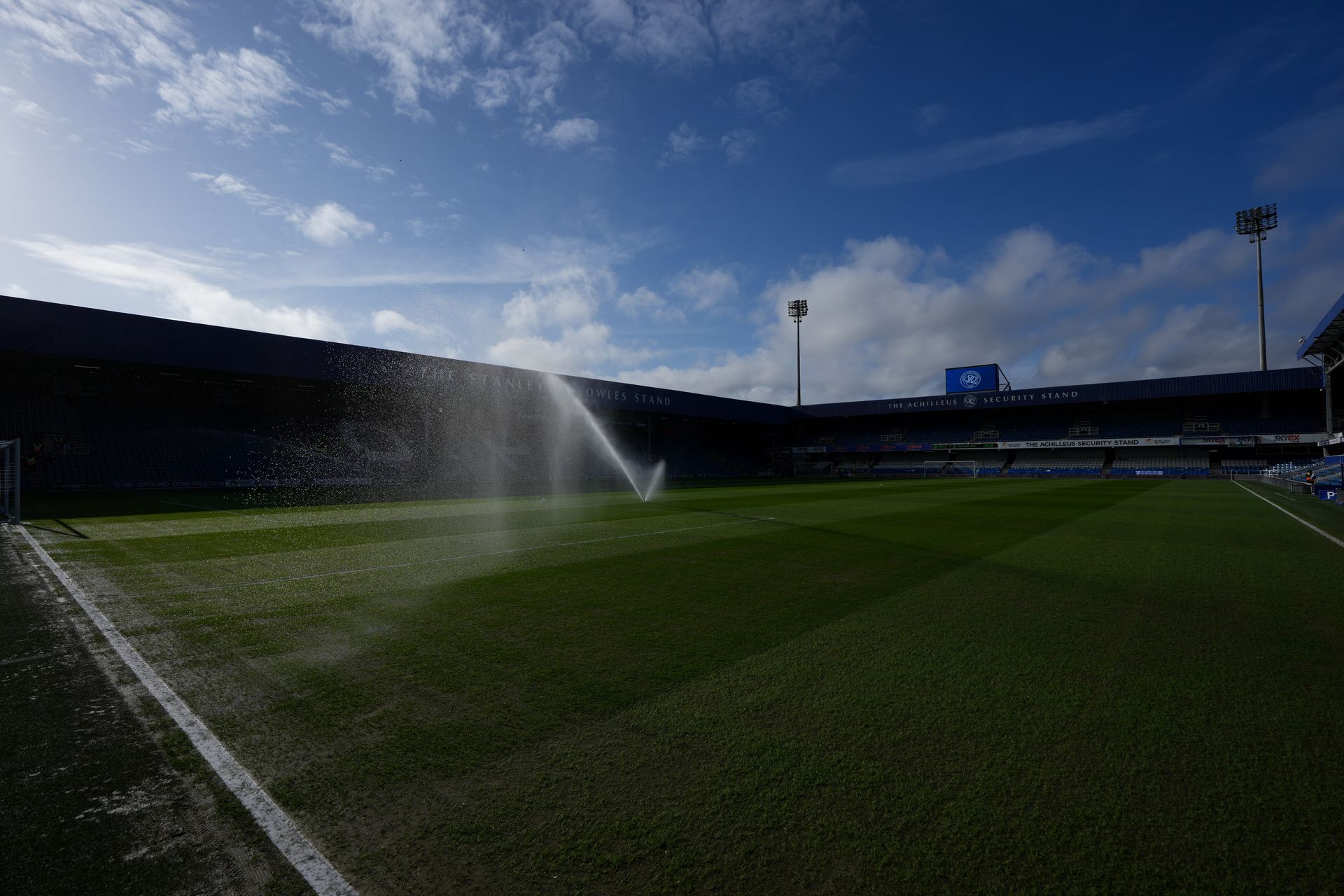 Flooded soccer field under blue sky with sprinklers spraying water across the pitch