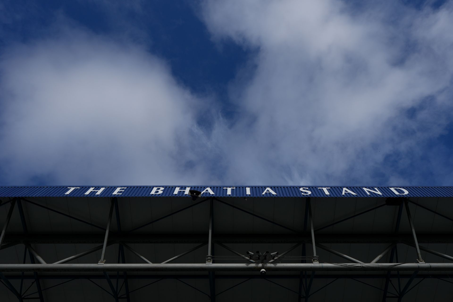 Upper facade of a stadium under a cloudy blue sky, with partial white signage along the roofline