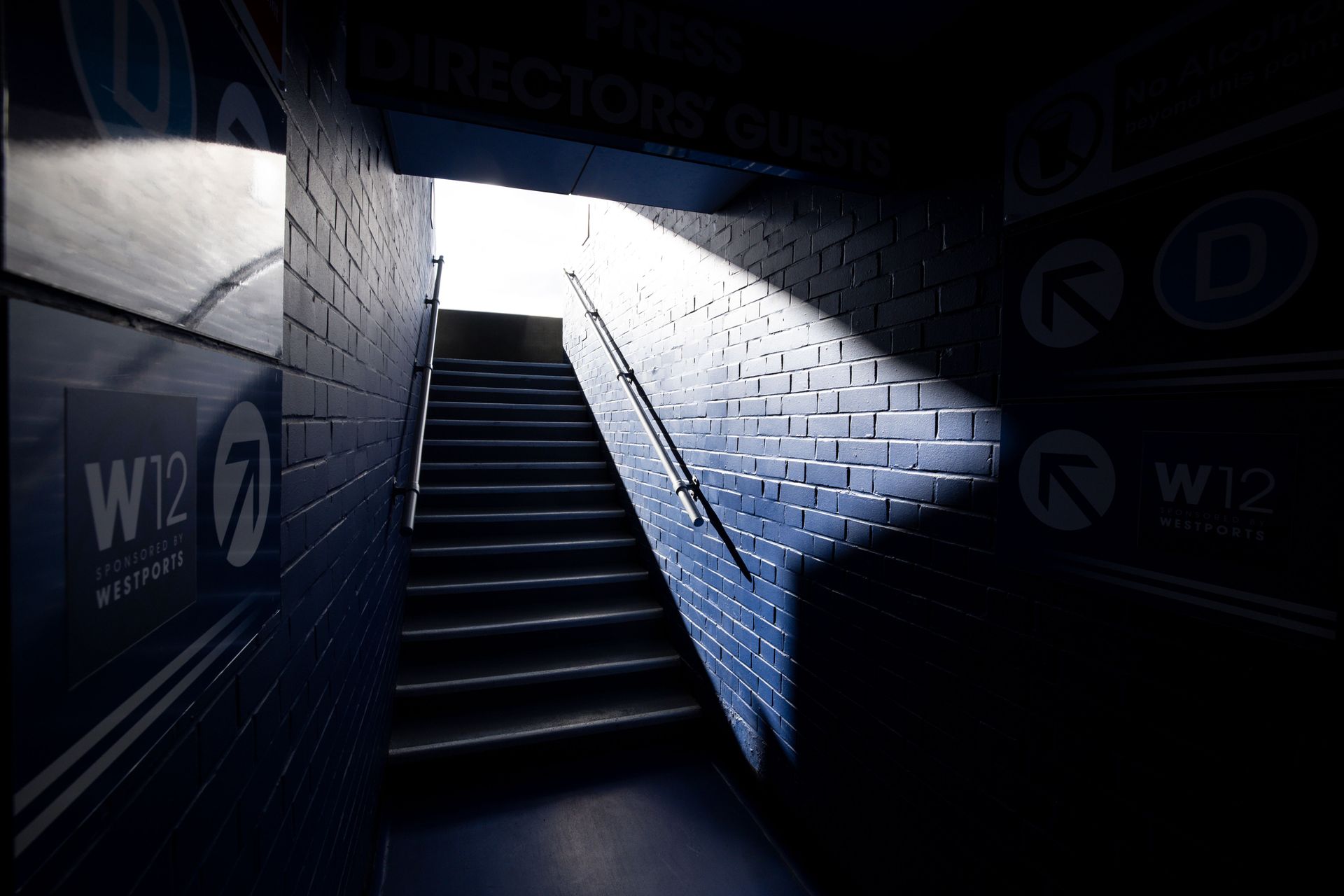 Dim stairwell lit by a bright spotlight, with blue-gray walls and shadows in a subway or underground passage